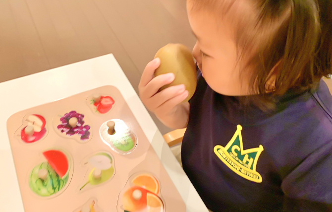 果物の型はめパズルと実際の果物キウイ。子どもがキウイの匂いを嗅いでいます。